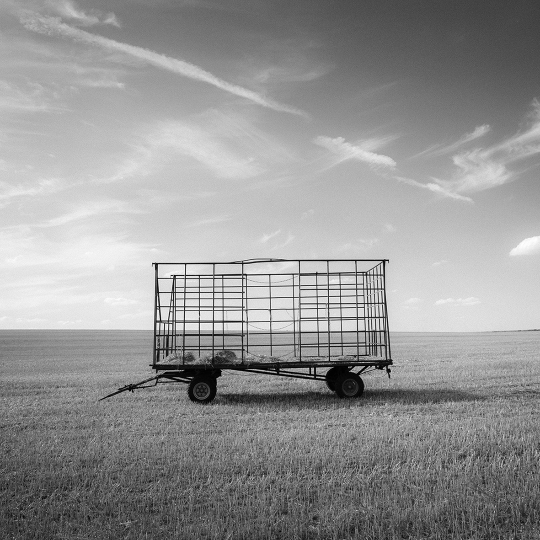 Ein mir Heuresten befüllter Heuwagen steht einsam und wie vergessen auf einem großen, menschenleeren und abgeernteten Acker. Am Himmel leichte Schleierwolken, der Blick geht bis zum Horizont.